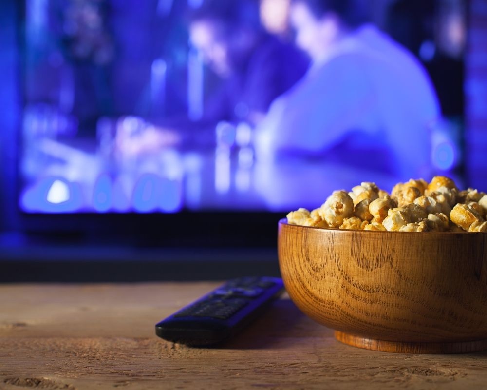 Primer plano de un fuente con palomitas, un mando a distancia y de fondo la televisión.
