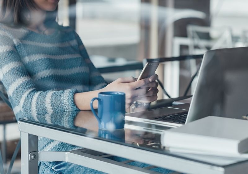 Imagen recortada de una chica escribiendo en su móvil y con ek portátil en su mesa de trabajo.
